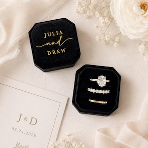 May include: A black velvet octagonal ring box with the gold text JULIA and DREW, containing three rings: an oval-cut solitaire, a diamond eternity band, and a plain gold band. Beside it lies a white card with the gold text J & D 05.24.2028.