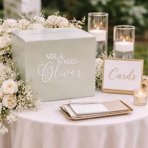 May include: A pale green card box with "Mr. & Mrs. Oliver" in white script is on a white tablecloth. A gold-framed "Cards" sign and candles are nearby. White flowers and greenery surround the box, creating a wedding theme.