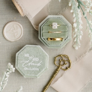 May include: A sage green velvet ring box with a white inscription that reads "Mr. & Mrs.  06.13.2020". The box is open and contains three gold rings, including a square-cut diamond engagement ring.