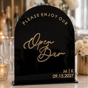 May include: An arched black acrylic sign featuring gold lettering that reads "PLEASE ENJOY OUR Open Bar" with the initials "M|K" and the date "09.15.2027" at the bottom. The sign is mounted on a rectangular black base and has a glossy finish.