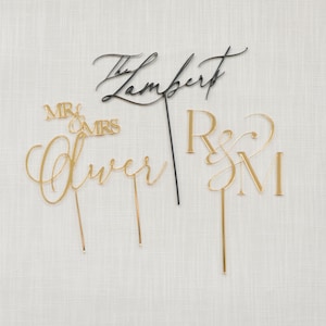 May include: A collection of elegant cake toppers in gold and black. The toppers feature cursive text with names and initials, including "Lambert," "Oliver," and "R&M," along with "Mr & Mrs." on a white background.