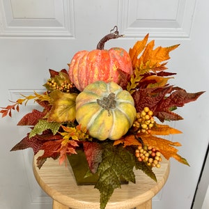Puede incluir: Un centro de mesa oto&ntilde;al con dos calabazas en tonos naranja y amarillo, rodeadas de coloridas hojas de oto&ntilde;o y bayas. El arreglo est&aacute; en un recipiente cuadrado de metal verde, creando una exhibici&oacute;n festiva para la temporada.