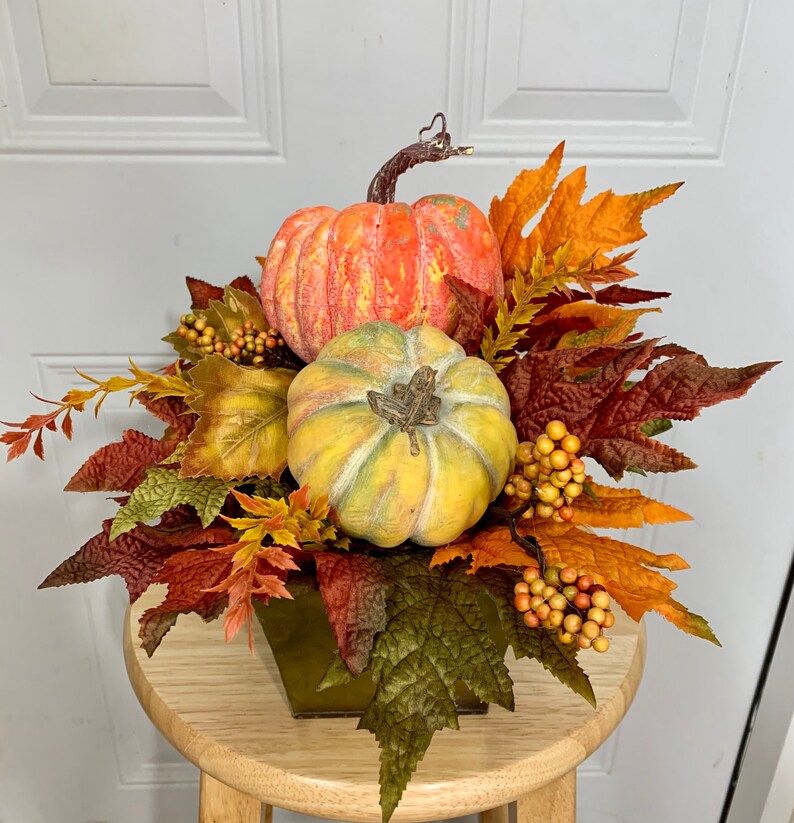 Puede incluir: Un centro de mesa oto&ntilde;al con dos calabazas en tonos naranja y amarillo, rodeadas de coloridas hojas de oto&ntilde;o y bayas. El arreglo est&aacute; en un recipiente cuadrado de metal verde, creando una exhibici&oacute;n festiva para la temporada.