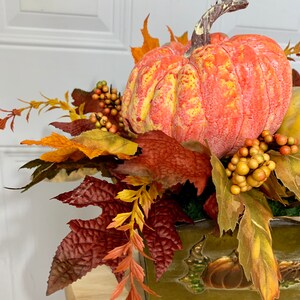 Puede incluir: Un centro de mesa con tem&aacute;tica oto&ntilde;al que presenta una gran calabaza naranja con detalles amarillos, rodeada de coloridas hojas y bayas de oto&ntilde;o. El arreglo est&aacute; en un recipiente verde decorativo con dise&ntilde;os de calabaza en relieve, ideal para la decoraci&oacute;n de temporada.
