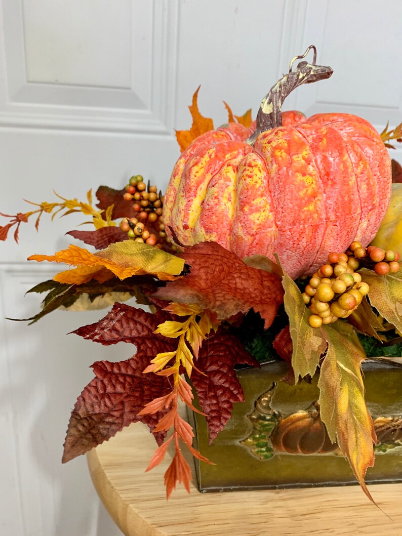 Puede incluir: Un centro de mesa con tem&aacute;tica oto&ntilde;al que presenta una gran calabaza naranja con detalles amarillos, rodeada de coloridas hojas y bayas de oto&ntilde;o. El arreglo est&aacute; en un recipiente verde decorativo con dise&ntilde;os de calabaza en relieve, ideal para la decoraci&oacute;n de temporada.
