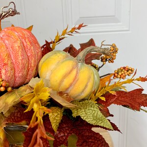 Puede incluir: Arreglo oto&ntilde;al con dos calabazas en tonos naranja y amarillo, rodeado de follaje artificial de oto&ntilde;o. El arreglo incluye hojas en rojo, verde y amarillo, junto con peque&ntilde;as bayas naranjas, creando un tema de cosecha.