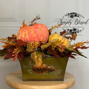 Puede incluir: Un centro de mesa con tem&aacute;tica oto&ntilde;al que presenta calabazas naranjas y amarillas, bayas y hojas de oto&ntilde;o dispuestas en un recipiente de metal verde. El recipiente tiene ilustraciones de calabazas. Un acento decorativo para la temporada de oto&ntilde;o.