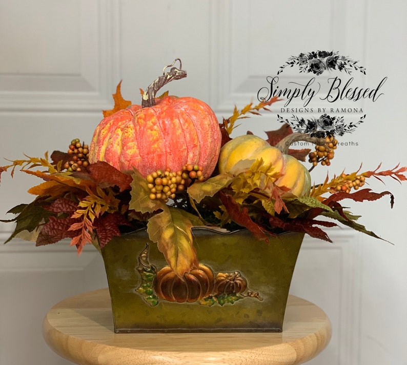 Puede incluir: Un centro de mesa con tem&aacute;tica oto&ntilde;al que presenta calabazas naranjas y amarillas, bayas y hojas de oto&ntilde;o dispuestas en un recipiente de metal verde. El recipiente tiene ilustraciones de calabazas. Un acento decorativo para la temporada de oto&ntilde;o.
