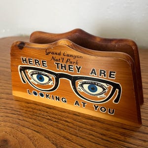 May include: Wooden napkin holder with the text "Grand Canyon Nat'l Park HERE THEY ARE LOOKING AT YOU." The holder features a graphic of eyeglasses with blue eyes. The wood has a natural grain and a warm brown color.
