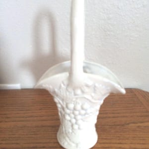 May include: White ceramic vase shaped like a basket with a handle. The vase is decorated with a grapevine design.