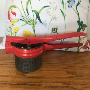 May include: Red metal potato ricer with a black mesh strainer. The ricer has a handle that is bent at a 90-degree angle.