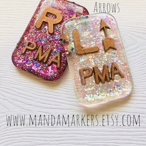 May include: Two rectangular resin keychains with rose gold letters and glitter. One is pink with the letter "R" and "PMA", the other is clear with the letter "L", an arrow, and "PMA". Text on top reads "Rose Gold Letters With Upright Arrows".