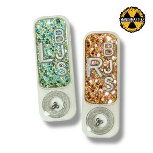 May include: Two white, rectangular accessories with glittery tops. One has teal and blue glitter with the letters "LJS", the other has gold and pink glitter with "RJS". Each has a silver circle with a small design.