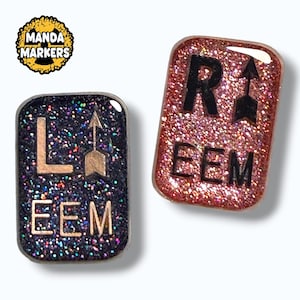 May include: Two rectangular pins with rounded corners. The left pin is dark purple with multicolored glitter and the text L, an arrow, and EEM. The right pin is rose gold with glitter and the text R, an arrow, and EEM. MANDA MARKERS logo is visible.