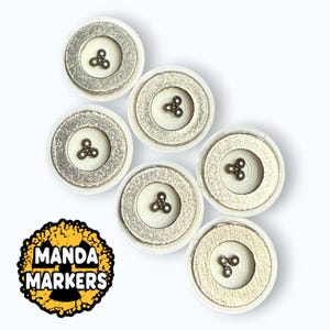 May include: A collection of six white and silver magnetic markers with a three-ball design in the center. The markers are arranged on a white background. The image also includes a logo that says "Manda Markers".