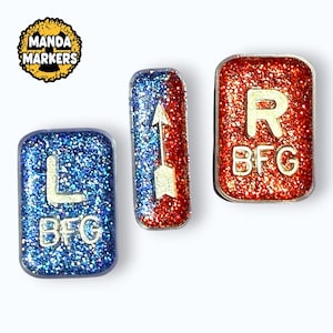 May include: Three glittery markers with rounded edges. One is blue with a white "L" and "BFG", another is red and blue with a white arrow, and the last is red with a white "R" and "BFG". The logo "Manda Markers" is in the upper left.