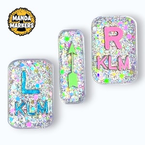 May include: Three glittery resin markers with the letters "L", "R", and an arrow. The letters and arrow are in bright colors, and the markers have the letters "KLM" at the bottom. The "Manda Markers" logo is in the upper left corner.