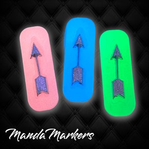 May include: Three rectangular markers in pink, blue, and green, each featuring a glittery arrow design. The markers are arranged against a black, textured background, with the text "Manda Markers" in white cursive at the bottom.