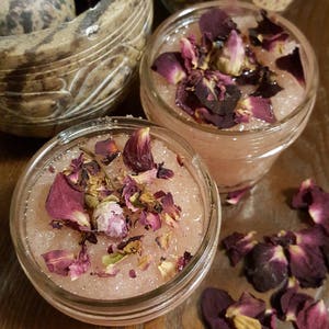 May include: Close-up of two small glass jars filled with a pink, granular substance and topped with dried rose petals. A mortar and pestle and a jar with a lid are in the background. The jars sit on a wooden surface.