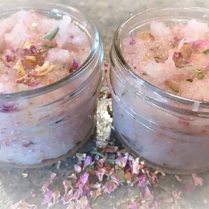 May include: Two small glass jars filled with pink, granular scrub. The scrub is speckled with dried rose petals and green herbs. The jars sit on a light gray surface, with scattered petals around them. The jars have a clear, transparent appearance.