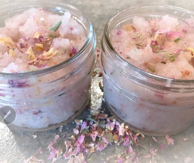 May include: Two small glass jars filled with pink, granular scrub. The scrub is speckled with dried rose petals and green herbs. The jars sit on a light gray surface, with scattered petals around them. The jars have a clear, transparent appearance.