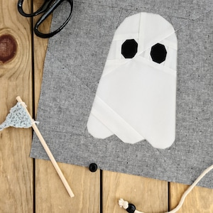 May include: A white ghost applique with black eyes on a gray fabric background. The ghost is made of fabric and has a simple, folded design.