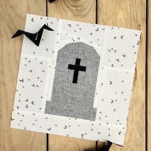 May include: A white fabric square with a black tombstone design in the center. The tombstone has a black cross on it. The fabric is decorated with a pattern of small black bats.