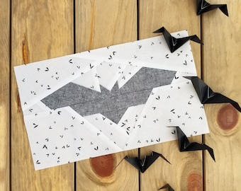 May include: A white card with a black bat design and a black and white pattern of small arrows. The card is surrounded by black origami bats on a wood background.