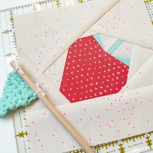 Puede incluir: Primer plano de un bloque de colcha de fresa de tela con fondo blanco y lunares rosas. La fresa es roja con corazones blancos y un tallo verde. Se puede ver una varilla de madera y una regla en el fondo.