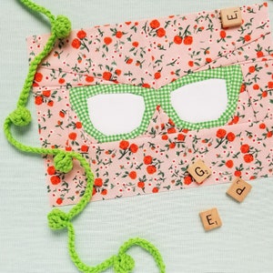 May include: A pink fabric panel with a green and white gingham pattern in the shape of glasses. The fabric panel is decorated with a floral print of small red roses and white flowers. The panel is surrounded by green yarn with small green pom-poms. The letters "E", "G2", and "E" are visible on the right side of the image.