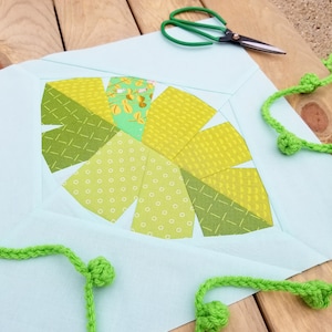 Pode incluir: Um quadrado de tecido azul claro com um design de patchwork verde e amarelo no centro. O design é um círculo com uma série de formas curvas que se assemelham a pétalas. O quadrado de tecido é rodeado por fio verde com pequenas bolas nas extremidades.