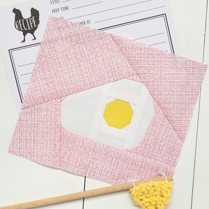 Pode incluir: Um quadrado de tecido rosa e branco com um círculo amarelo no centro. O quadrado de tecido é um bloco de colcha para um padrão de "Ovo Frito". Uma vareta de madeira com um triângulo de crochê amarelo está em primeiro plano.