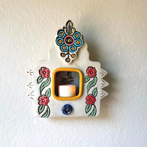 May include: A white ceramic wall-mounted candle holder featuring a central yellow-rimmed square opening containing a small white tea light candle. The piece is decorated with hand-painted red flowers, green vines, and a blue floral top accent.