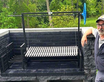 Op de afbeelding: Een zwarte buitenbarbecue met een metalen rooster en een stenen basis. De barbecue is opgezet in een achtertuin met een persoon ernaast.