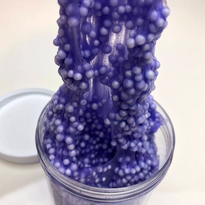 Può includere: Un barattolo di plastica trasparente pieno di slime viola, mescolato con piccole palline di schiuma bianche. Lo slime viene tirato fuori dal barattolo, mostrando la sua consistenza elastica. Il coperchio bianco del barattolo è accanto.
