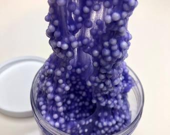 Könnte beinhalten: Ein durchsichtiges Plastikglas gefüllt mit lila Schleim, gemischt mit kleinen weißen Schaumstoffkügelchen. Der Schleim wird aus dem Glas gezogen und zeigt seine dehnbare Textur. Der weiße Deckel des Glases liegt daneben.