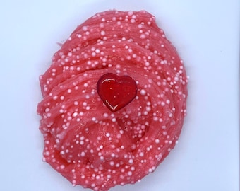 Könnte beinhalten: Nahaufnahme von rosa Schleim mit weißen Schaumperlen und einem herzförmigen roten Anhänger. Der Schleim hat eine leuchtend rosa Farbe, und die Perlen sorgen für Textur. Der Herzanhänger ist glänzend.