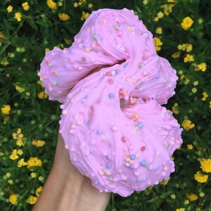 May include: Pink slime with colorful sprinkles, held in a hand against a background of green foliage with yellow flowers.