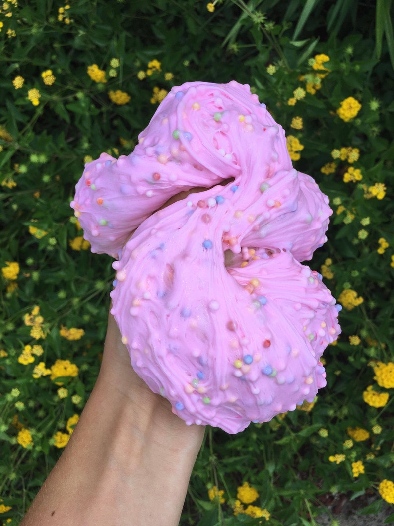 May include: Pink slime with colorful sprinkles, held in a hand against a background of green foliage with yellow flowers.