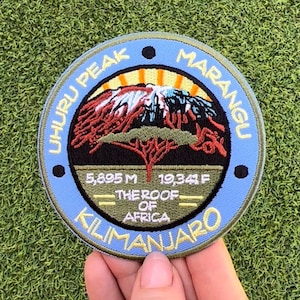 Könnte beinhalten: Ein blauer und grüner gestickter Aufnäher mit den Worten "Uhuru Peak" und "Marangu" auf einem Kreis. Der Aufnäher zeigt auch einen Berg mit Sonnenaufgang und den Text "5.895 m 19.341 ft - Das Dach Afrikas - Kilimanjaro".