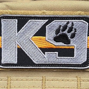 Könnte beinhalten: Ein grauer und schwarzer gestickter Patch mit den Buchstaben "K9" und einem Pfotenabdruck. Der Patch hat einen dünnen orangefarbenen Streifen oben.