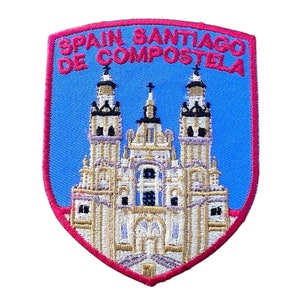 May include: Embroidered patch with a blue background and red border. The patch features a white building with gold accents and the text "Spain Santiago de Compostela".