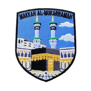 Puede incluir: Un parche bordado que representa la Kaaba en La Meca, Arabia Saudita. El parche presenta un fondo azul con nubes blancas, dos minaretes y el texto "Makkah Al-Mukarramah".