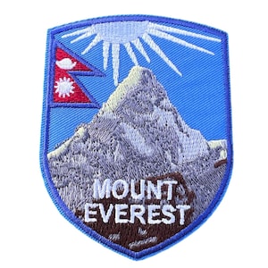 Può includere: Una toppa ricamata blu e bianca con una montagna e il testo "MOUNT EVEREST". La toppa presenta un sole bianco con raggi e una bandiera rossa e blu del Nepal.