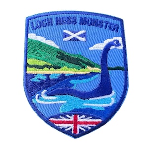Pode incluir: Etiqueta bordada azul e branca com o texto "Loch Ness Monster" e uma imagem do monstro do Lago Ness na água. A etiqueta também apresenta a bandeira escocesa e a Union Jack.