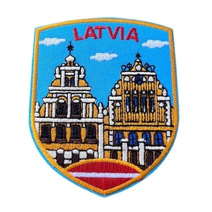 May include: Embroidered patch featuring a blue shield with a yellow and red Latvian flag at the bottom. The shield depicts two yellow buildings with red roofs against a blue sky with white clouds. The text "LATVIA" is embroidered in red at the top of the shield.
