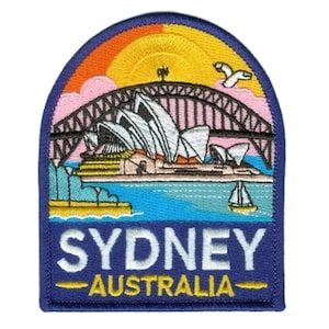 Może przedstawiać: Haftowana naszywka przedstawiająca Operę w Sydney i Most Portowy na tle zachodzącego słońca. Naszywka z niebieską obwódką zawiera napis "SYDNEY AUSTRALIA" w kolorze białym i żółtym.