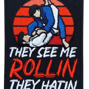Può includere: Toppa ricamata nera con una scena di arti marziali con due figure in uniforme blu e bianca contro un design tramonto rosso e arancione. Testo: "THEY SEE ME ROLLIN THEY HATIN" in bianco e rosso.