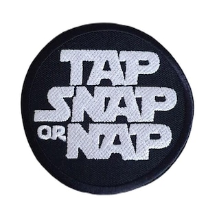 Könnte beinhalten: Schwarzer und weißer gestickter Aufnäher mit dem Text "Tap Snap Or Nap" in runder Form.