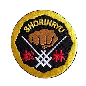 May include: A black and yellow embroidered patch with the text "Shorinryu" in a circle. The patch features a brown fist, two white sticks, and the text "松林" in red.